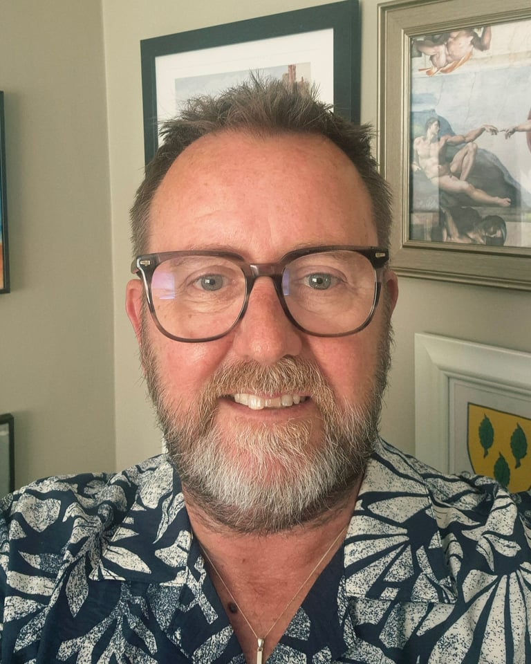 Middle-aged man with beard and glasses wearing a patterned blue shirt, smiling at the camera indoors